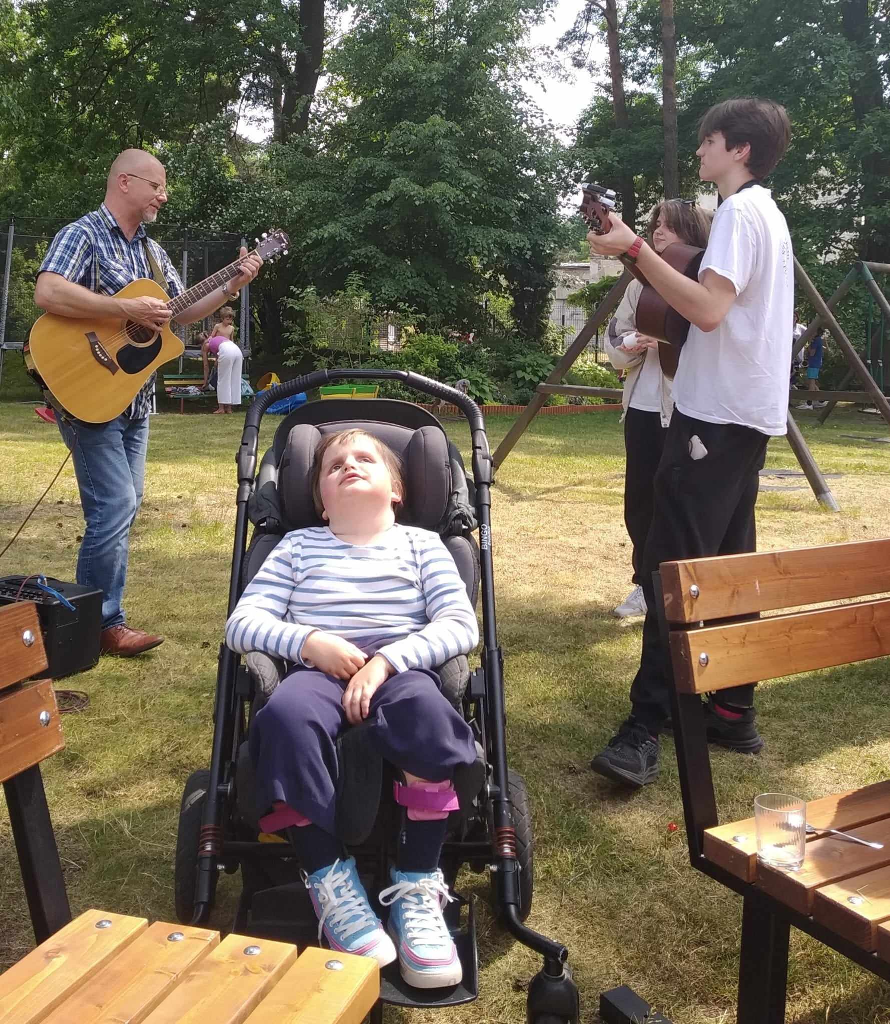 Dziewczynka siedzi na wózku, za nią chłopiec i jeden z ojców grają na gitarach.