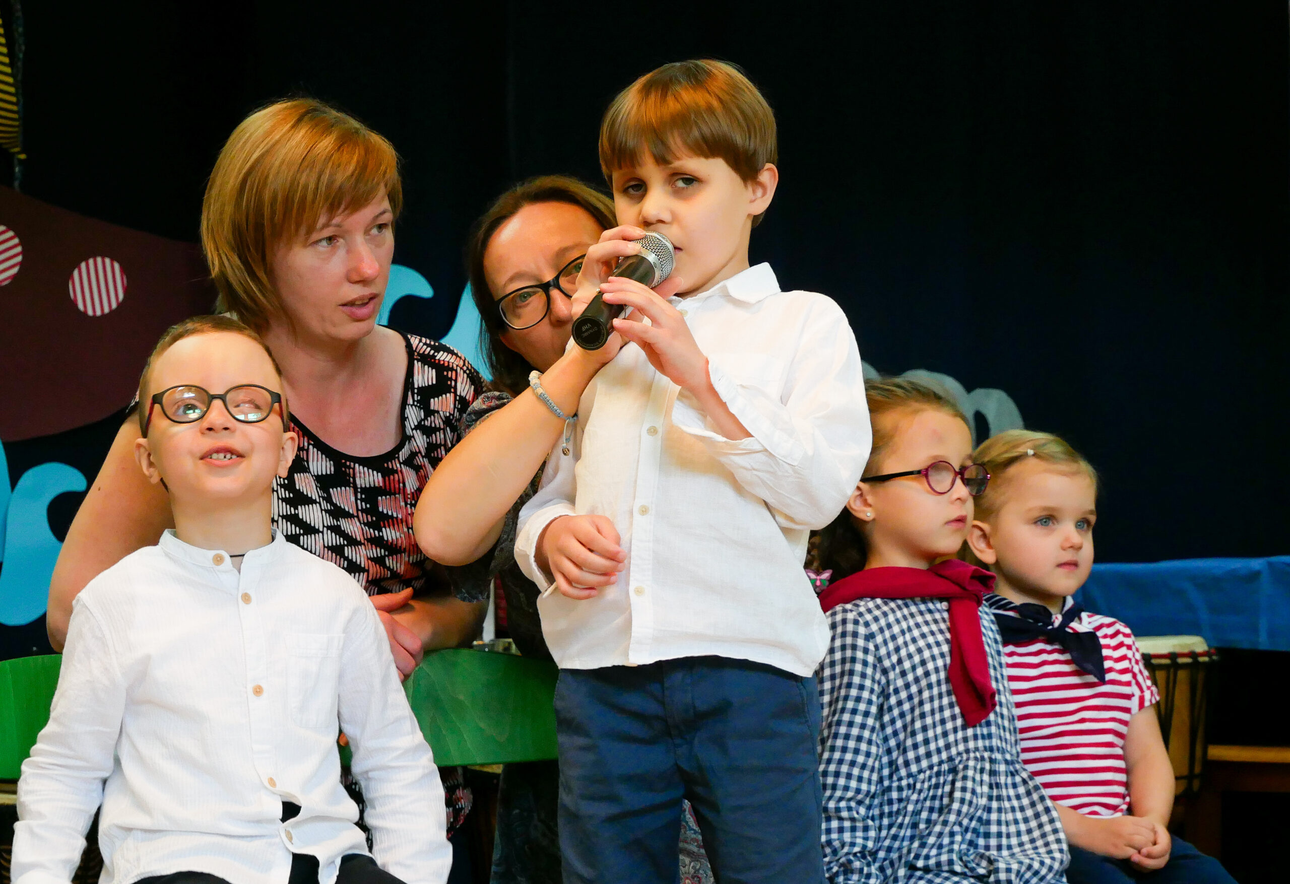 Chłopiec występuje na scenie z mikrofonem. Obok niego siedzi nauczycielka.