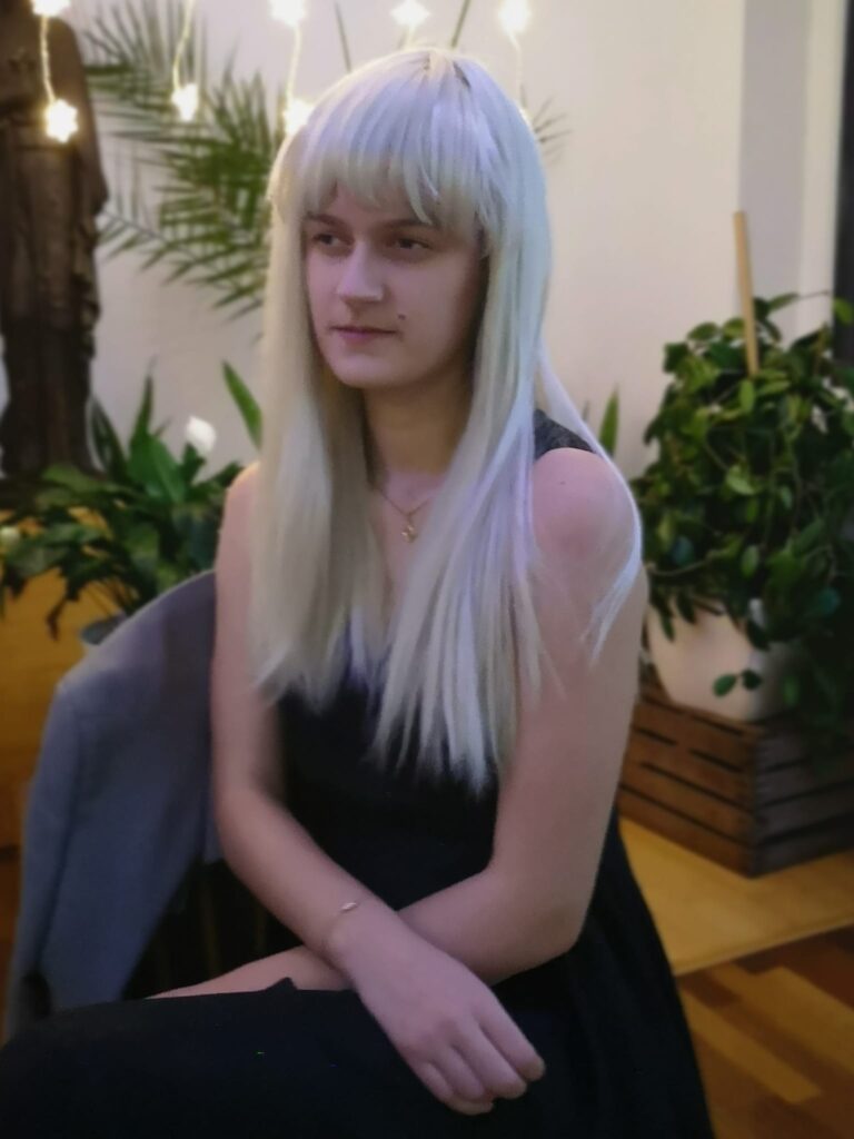 Karolina ubrana w czarną sukienkę a na głowie ma blond perukę, siedzi zamyślona