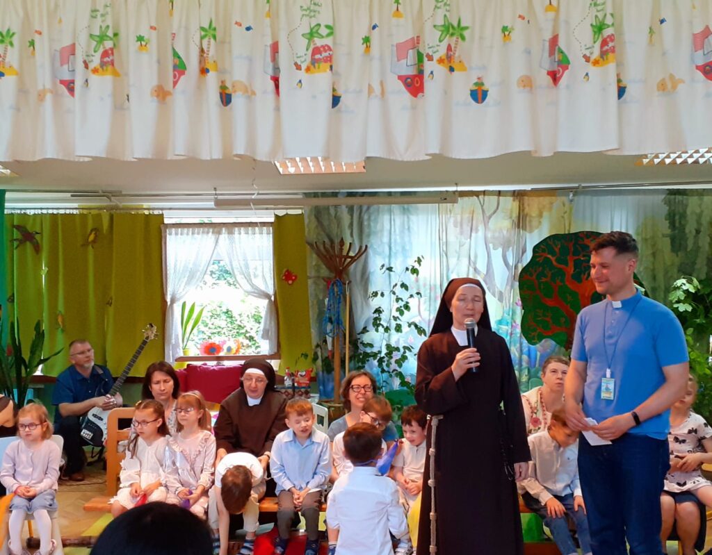 Siostra Hiacynta mówi przez mikrofon, ksiądz Sebastian stoi obok a w tle przedszkolaki siedzą na ławeczkach