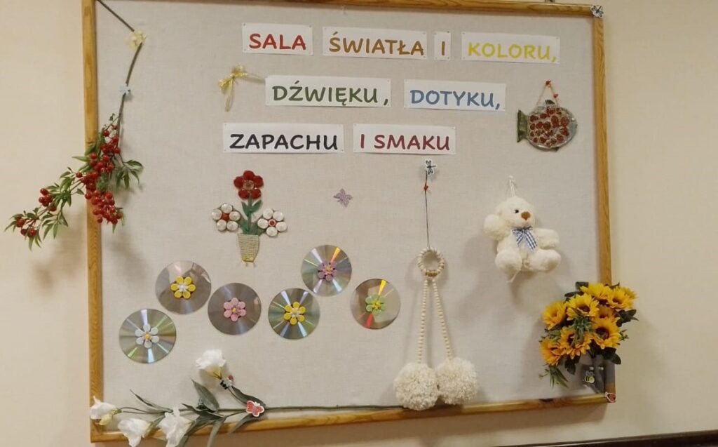 Na białej ścianie wisi drewniana ramka informująca o sali światła, koloru, dźwięku, dotyku, zapachu, smaku. 