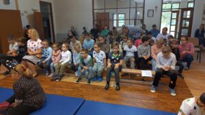 Uczniowie szkoły podstawowej siedzą na ławkach na korytarzu i słuchają przedstawienia.