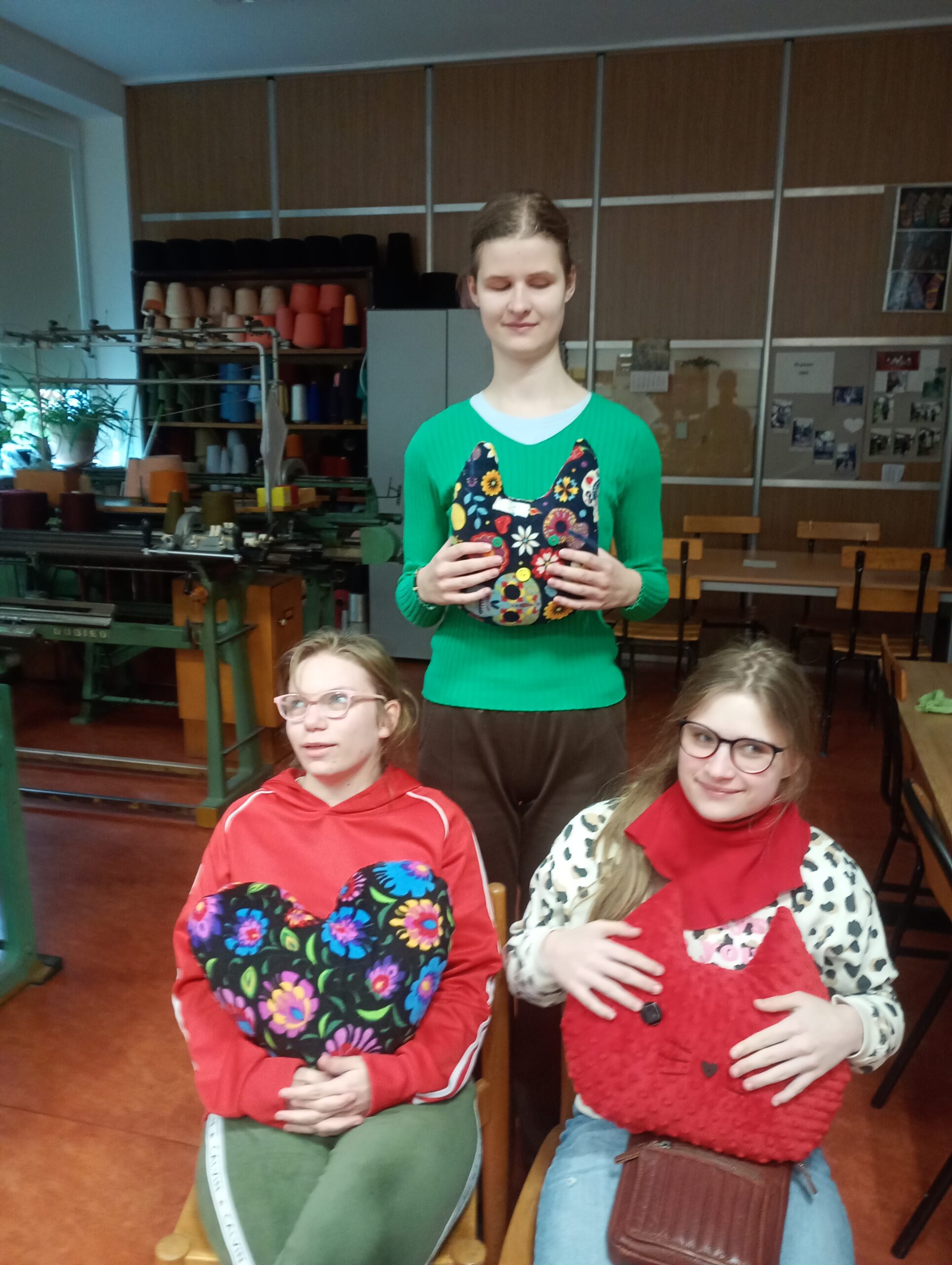 Trzy uczennice prezentują swoje poduszki. Jedna stoi z tyłu a dwie siedzą na krzesłach.