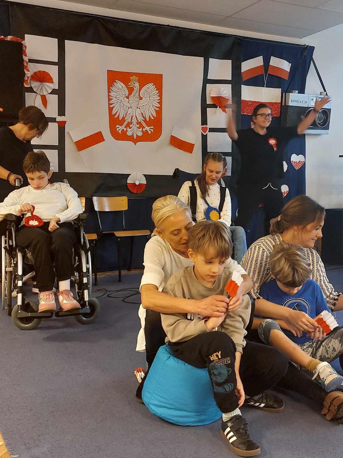 uczniowie klasy drugiej i klasy piatej z nauczycielami wykonują pieśń patriotyczną na szkolnej scenie