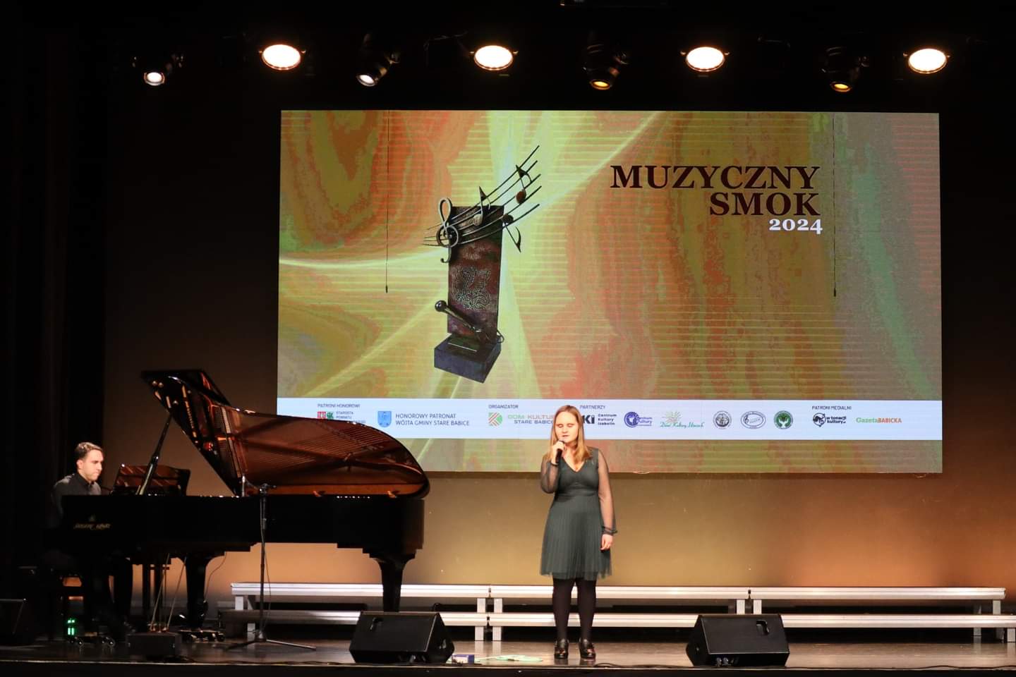 Julia Krynicka śpiewa na scenie, akompaniuje przy fortepianie Grzegorz Bożewicz
