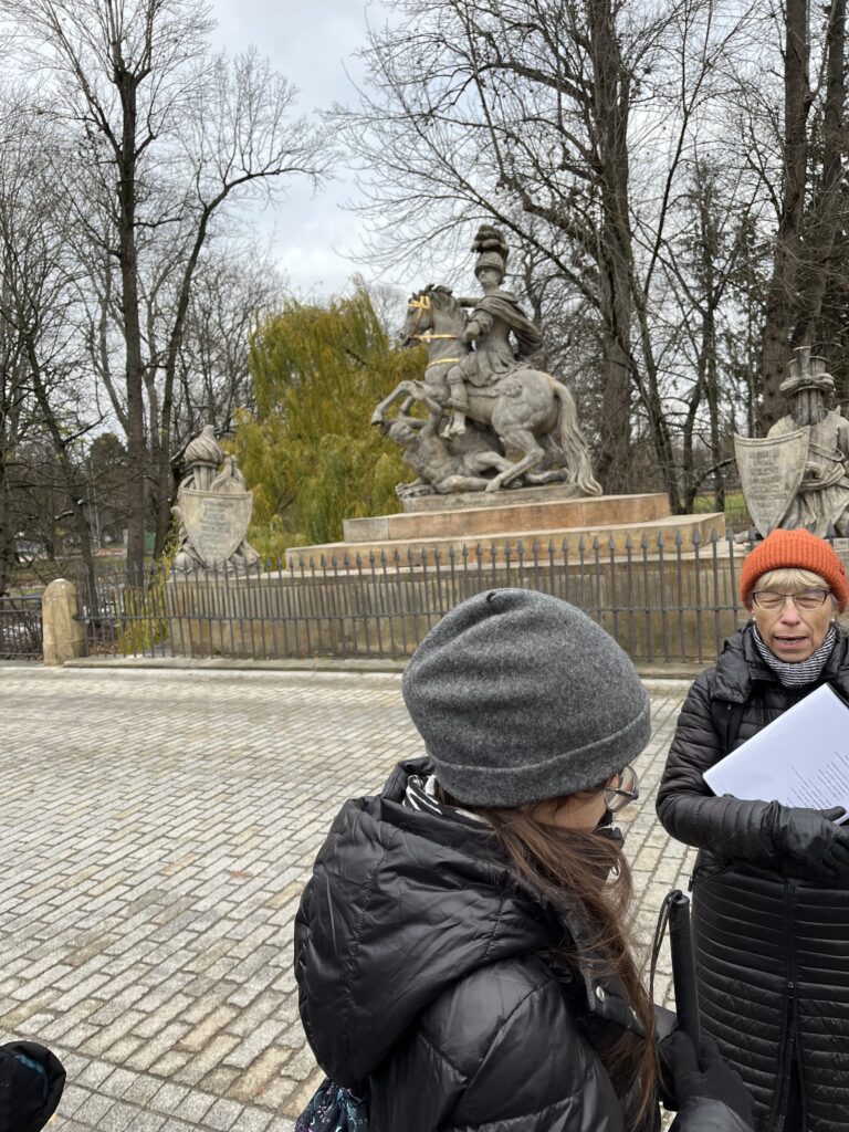 Uczennica wraz z nauczycielami przed pomnikiem z bohaterem na koniu.