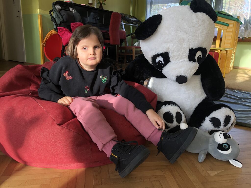 Dziewczynka siedzi na pufie sako obok wielkiej pluszowej pandy.