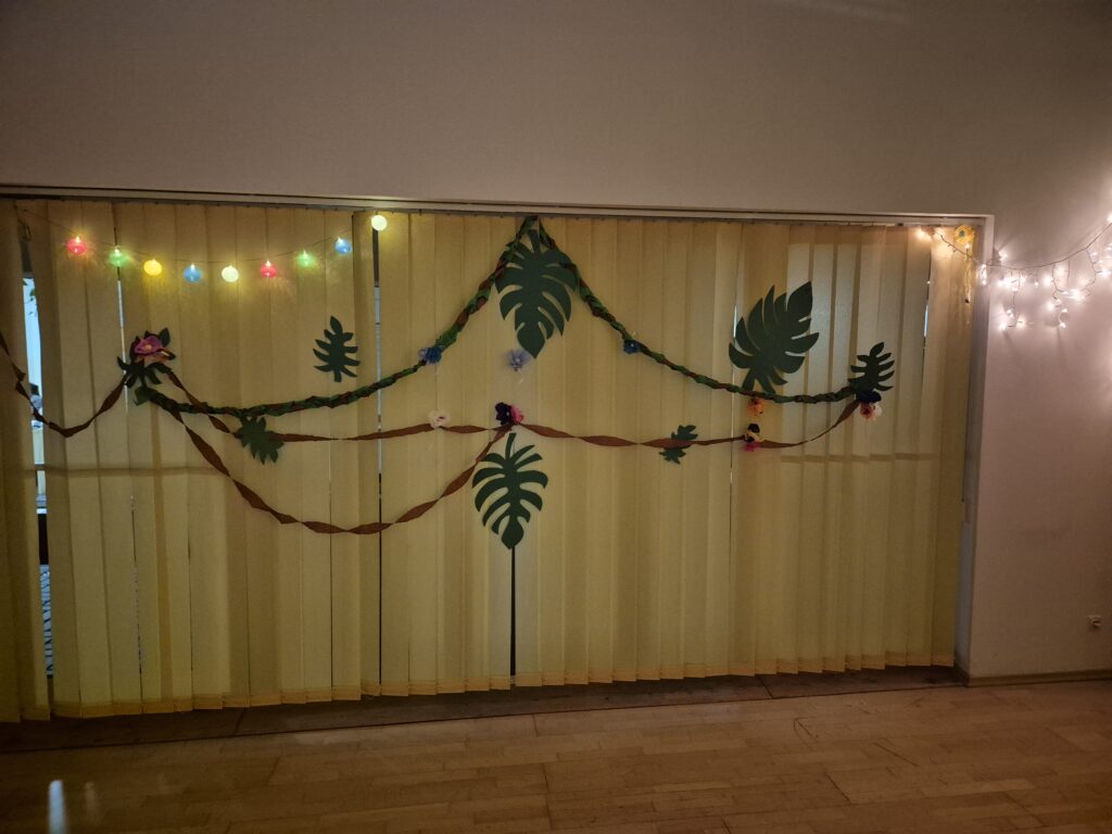 Na ścianie z oknami na wertikalach powieszone dekoracje hawajskie liście, liany, kwiaty