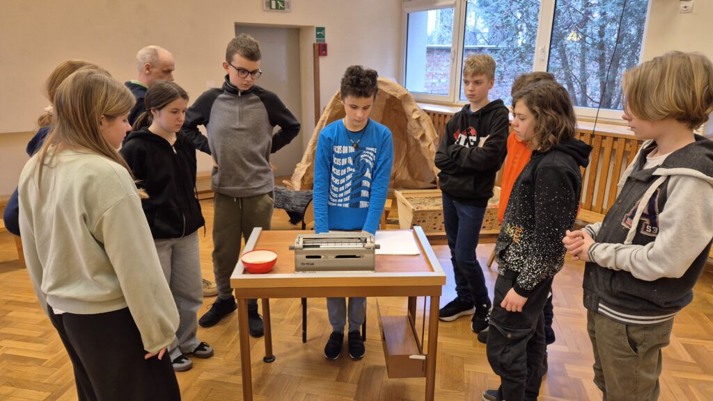 Uczniowie 7 klasy i 6 klasy na sali obserwują maszynę brajlowską.