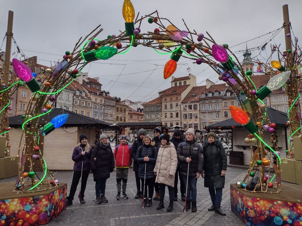 Uczniowie wraz z nauczycielami stoją pod świątecznym łukiem ozdobionym kolorowymi lampkami w kształcie ogromnych żarówek i bombek. W tle widać zabytkowe budynki Starego Miasta, które dodają uroku miejscu.