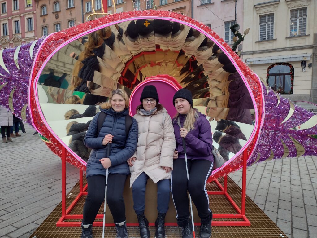 Trzy uczennice siedzą na ławce umieszczonej wewnątrz kolorowej, świątecznej instalacji w kształcie serca. Konstrukcja jest ozdobiona fioletowymi i czerwonymi elementami oraz lustrzanymi panelami, które tworzą ciekawy efekt odbicia. Osoby na zdjęciu są zadowolone i uśmiechnięte.