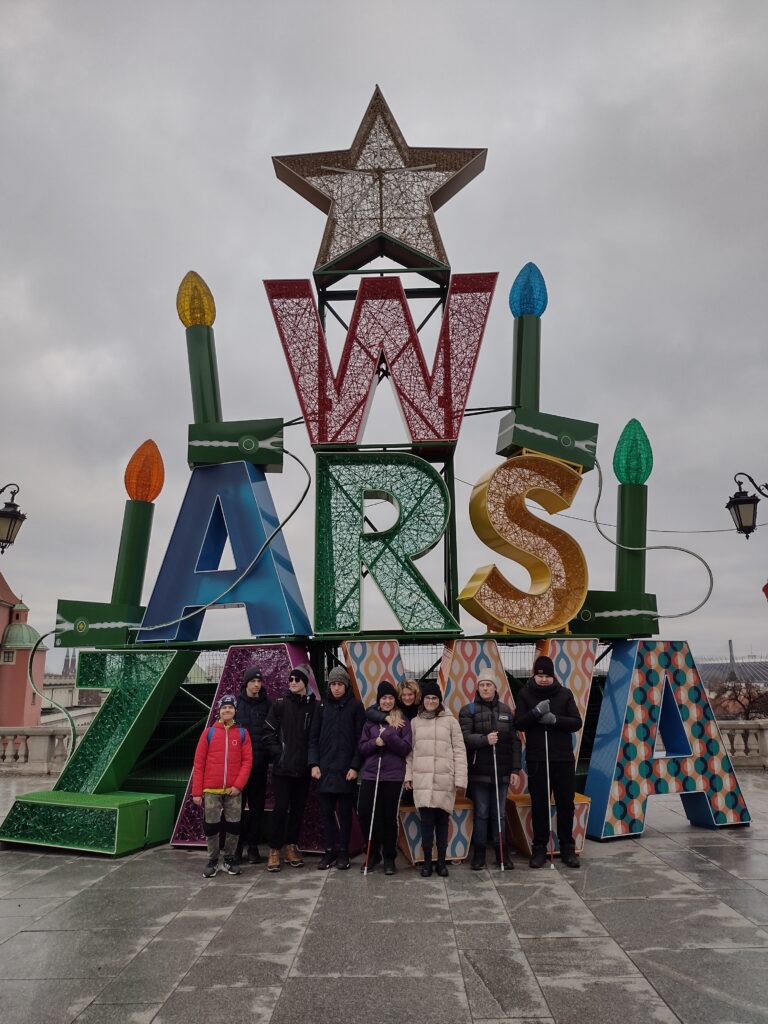Grupa stoi przed dużą, kolorową instalacją napisu "WARSZAWA", ozdobionego żarówkami i dużą gwiazdą na szczycie. Instalacja ma świąteczny charakter.