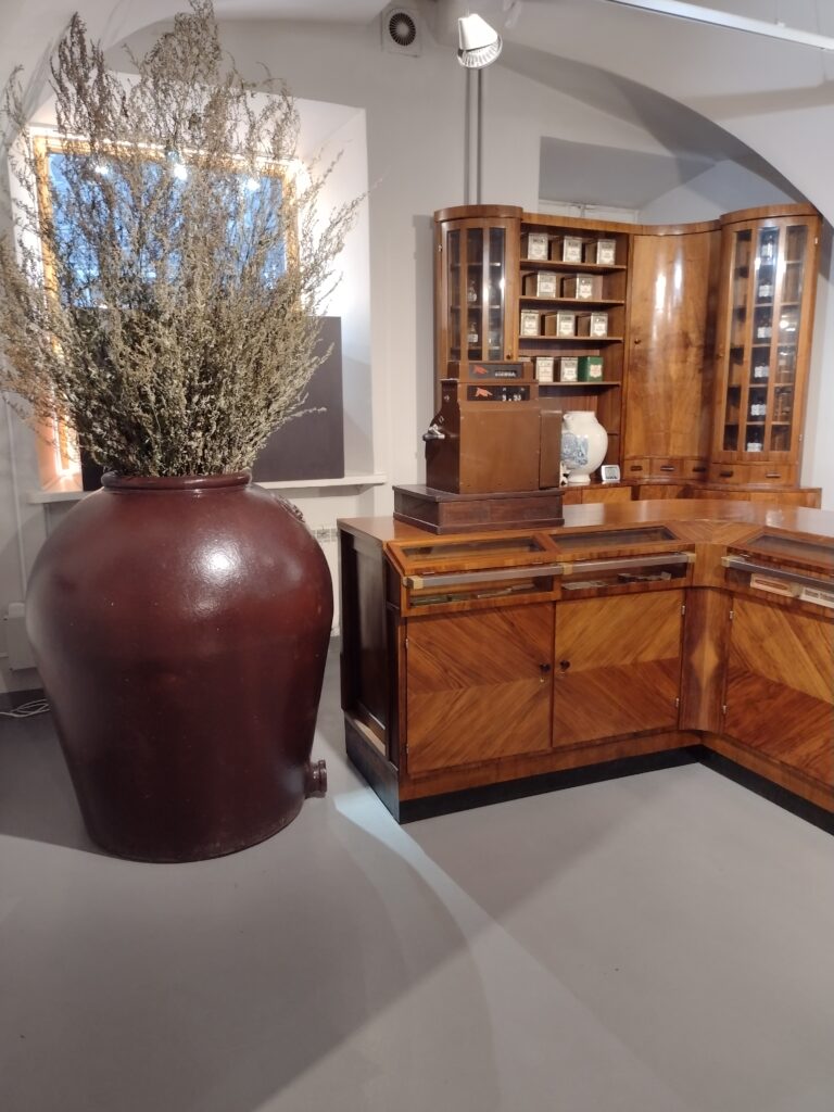 Wnętrze muzeum farmacji . Po lewej duży wazon z suszonymi kwiatami po prawej drewniane biurko oraz szafki z lekarstwami.