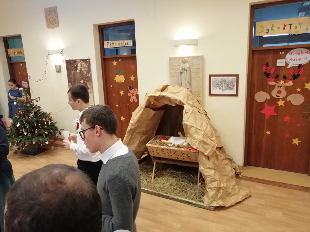 Żłobek Jezusa w tle. Uczniowie z opłatkami słuchają życzeń Pani dyrektor.