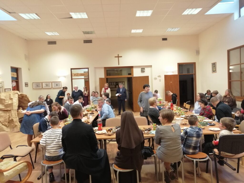 Uczestnicy słuchają śpiewanej kolędy. Siedzą radośnie przy stołach z potrawami. 