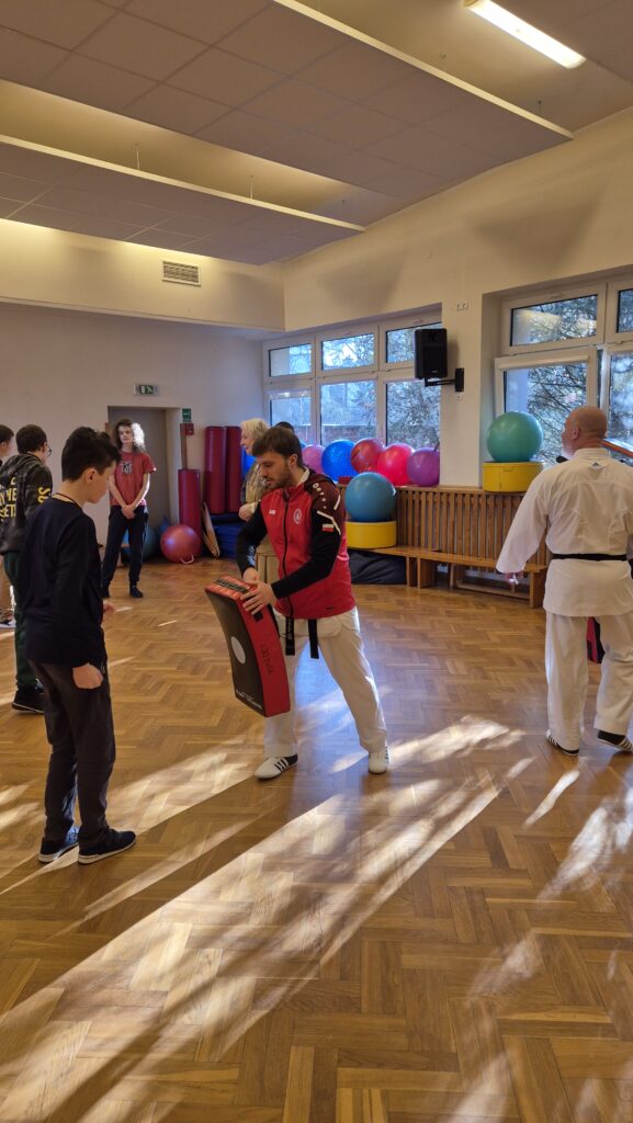 Na sali gimnastycznej trenerzy pokazują podstawowe chwyty karate. Na pierwszym planie uczeń przygotowuję się do ataku.