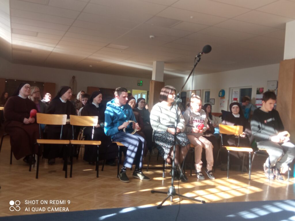 Siostry i uczniowie siedzą na krzesłach i śpiewają