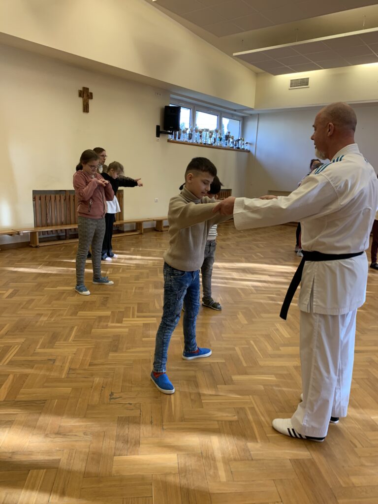 Na sali gimnastycznej trener trzyma dłońmi dłonie ucznia prezentują chwyt karate. W tle uczniowie słuchają.