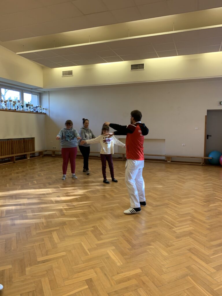 Na sali gimnastycznej trener pokazuje dłońmi chwyt karate. Uczennice naśladują ruchy i uważnie słuchają poleceń trenera.