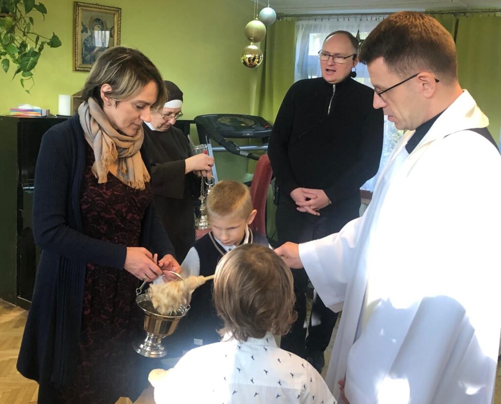 Wychowawczyni wraz z chłopcami asystują księdzu, w czasie poświęcania bawialni
