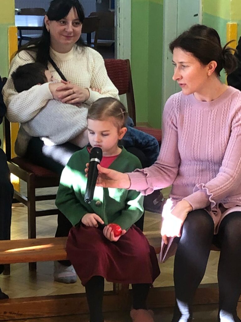 Mama z chłopcem śpiącym na kolanach, dziewczynka siedząca na ławce obok pani trzyma mikrofon przy jej twarzy