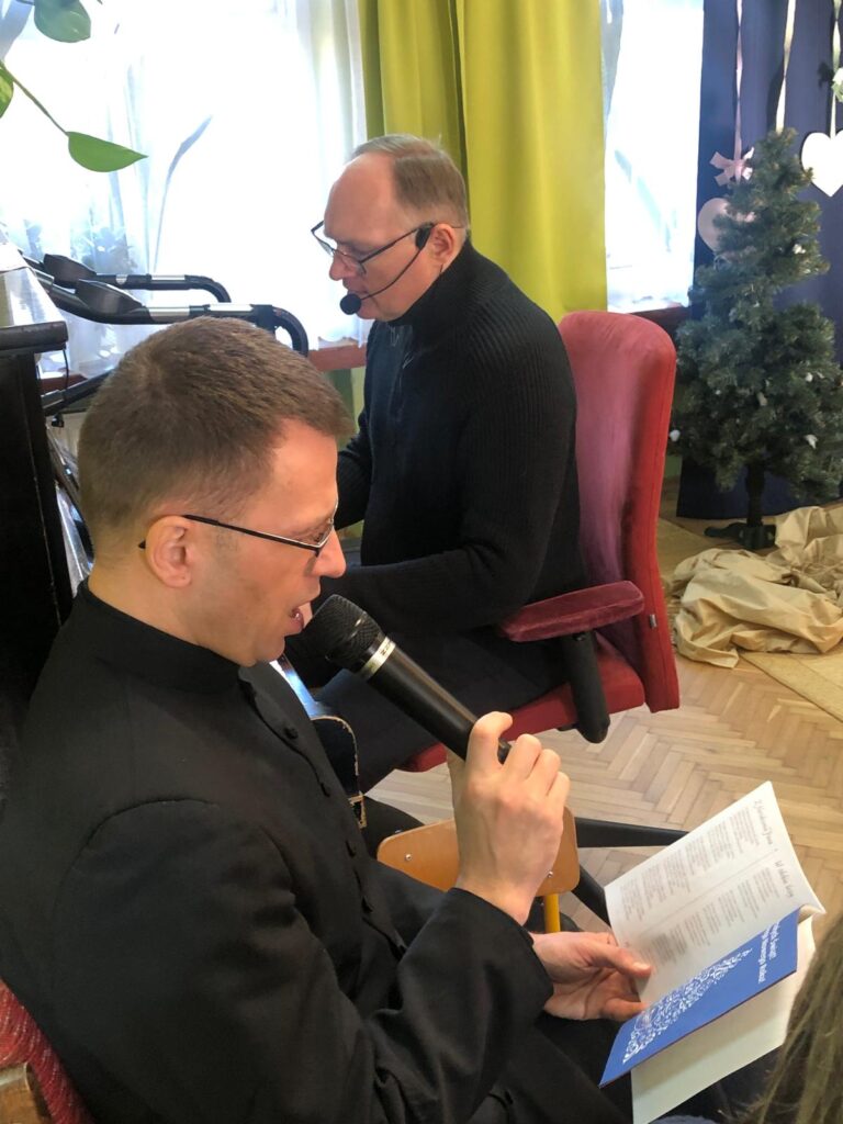 Pan Tomek gra na pianinie obok siedzi ksiądz Marek z mikrofonem i śpiewnikiem i śpiewa