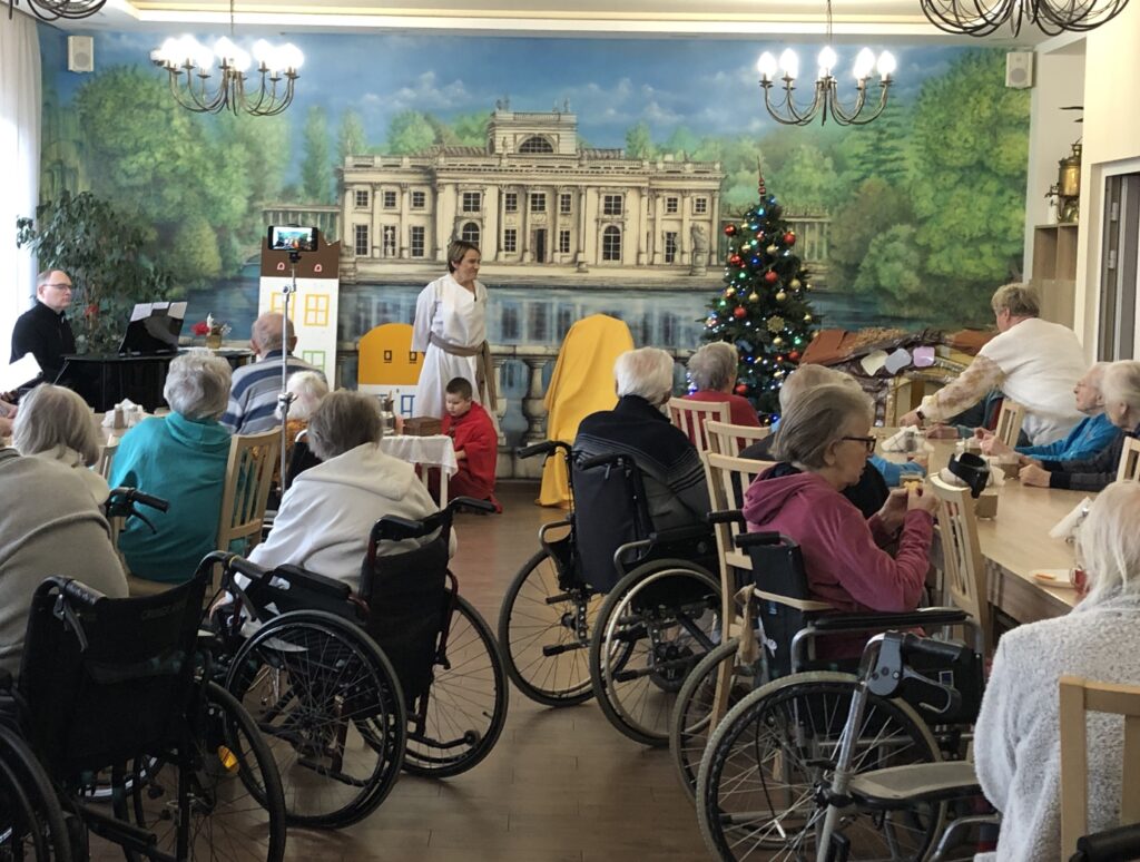 Seniorzy siedzący na wózkach inwalidzkich patrzą w stronę sceny, na której znajduje się chłopiec w czerwonym płaszczu i nauczycielka w białej szacie. 
