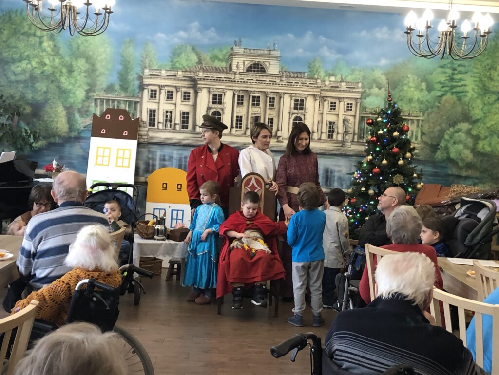 Na scenie grupa dzieci i nauczycieli w przebraniach teatralnych szykują się do śpiewania kolędy. Po bokach seniorzy siedzą na wózkach inwalidzkich i patrzą na aktorów.