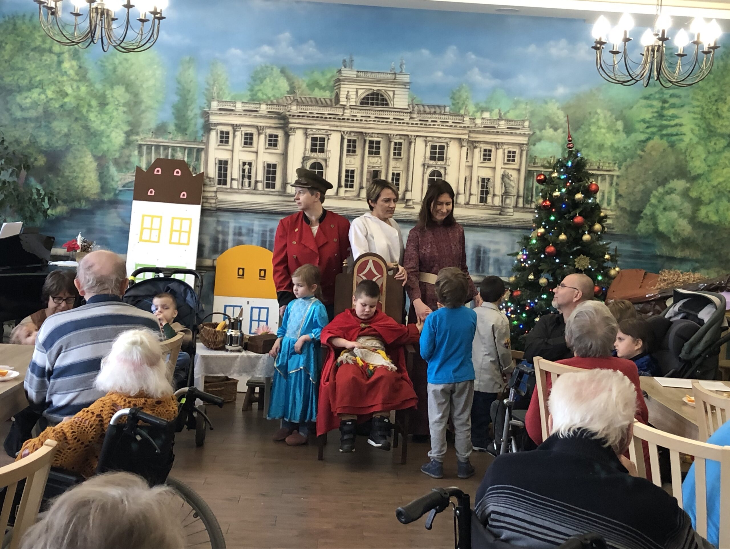 Na scenie grupa dzieci i nauczycieli w przebraniach teatralnych szykują się do śpiewania kolędy. Po bokach seniorzy siedzą na wózkach inwalidzkich i patrzą na aktorów.