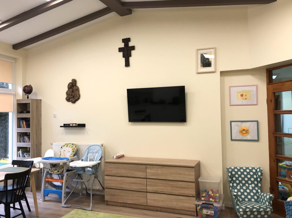 Na zdjęciu widać komodę z szufladami a nad nią duży telewizor. Po lewej stronie stoją regał z książkami i krzesełka do karmienia małych dzieci. Po prawej stronie pojemniki z klockami i zielony fotel w grochy dla dzieci. Całość uzupełnia dekoracja na ścianie: płaskorzeźba Świętej Rodziny, drewniany krzyż, zdjęcie Bł. Elżbiety Róży Czackiej.
