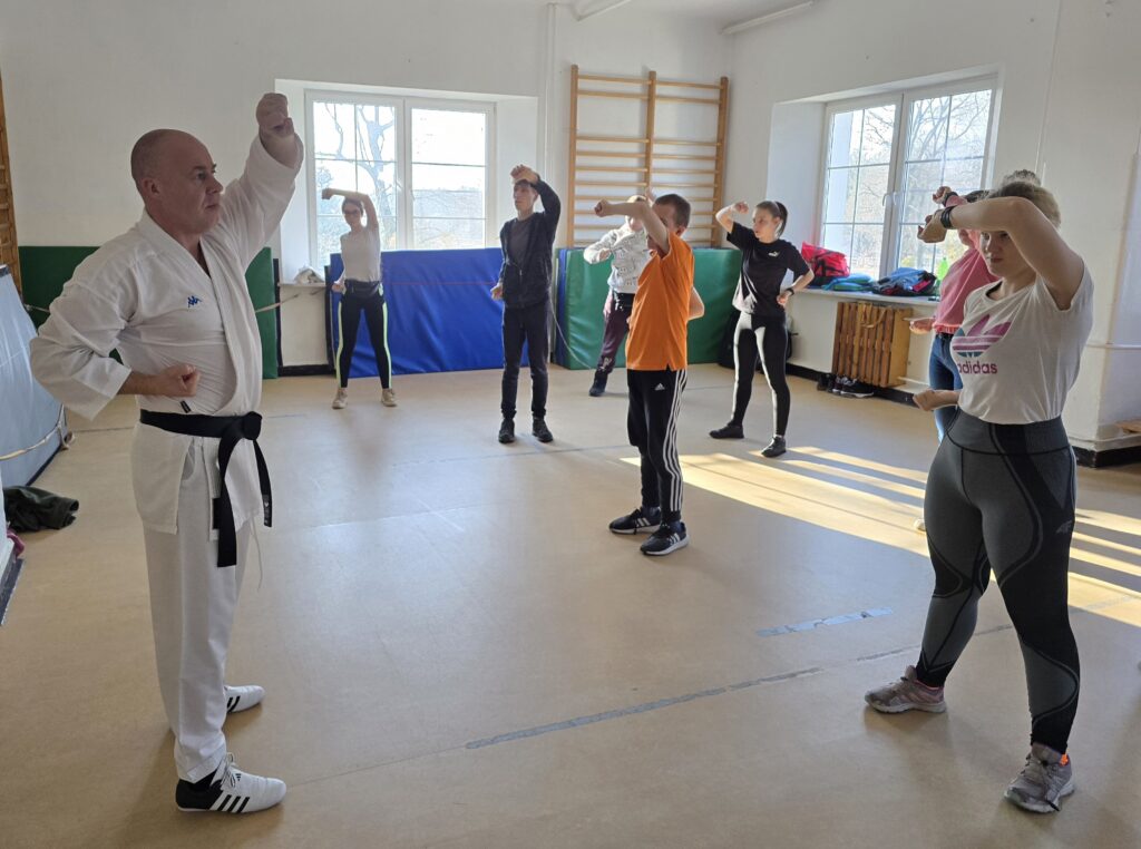 Trener pokazuje podstawowe chwyty karate na sali gimnastycznej. Uczniowie naśladują ruchy dłońmi trenera stojąc i kierując dłonie w celu samoobrony.
