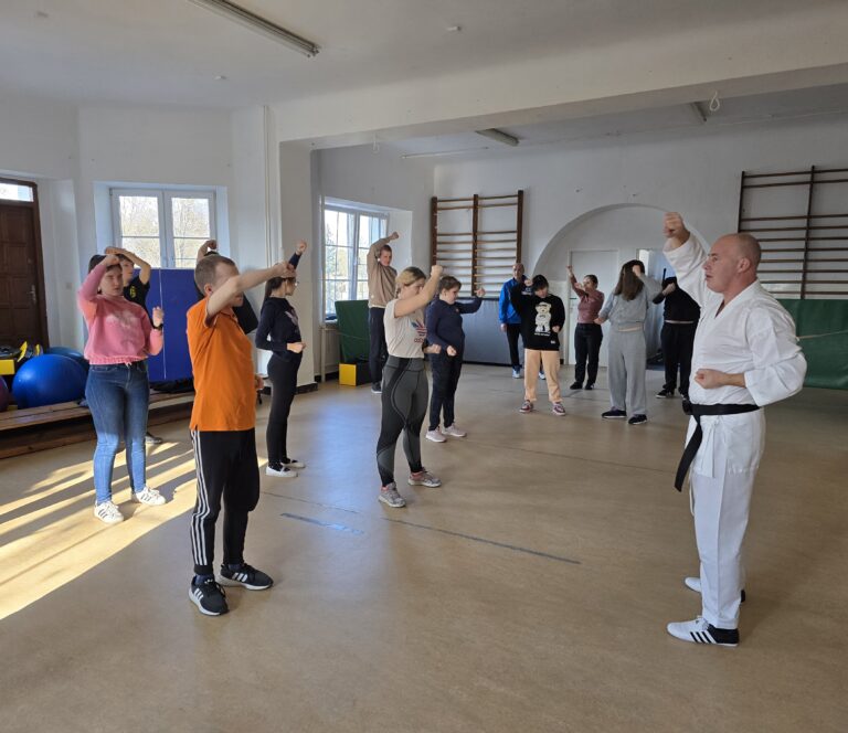 Trenerzy pokazują podstawowe chwyty karate na sali gimnastycznej. Uczniowie naśladują ruchy dłońmi trenera stojąc i słuchając wypowiedzi.