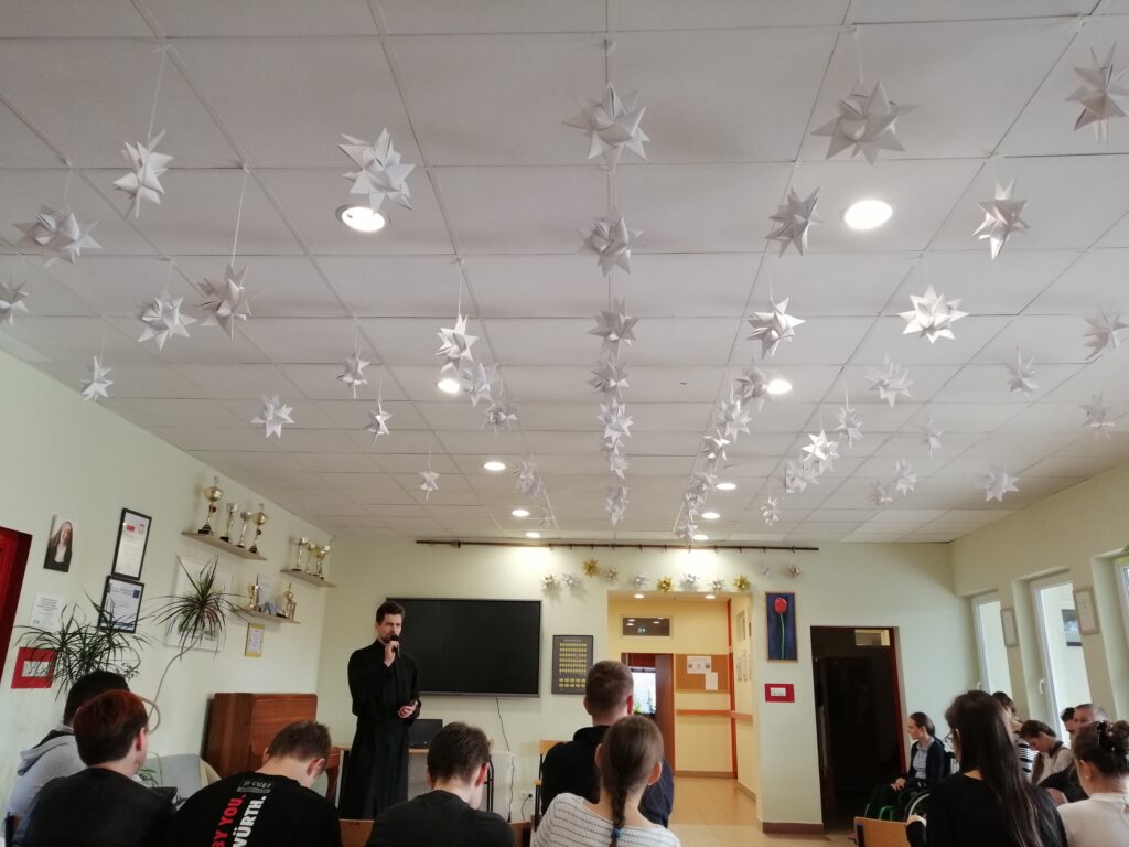 Uczniowie zgromadzeni na korytarzu siedząc na krzesłach słuchają wypowiedzi brata Krzysztofa.