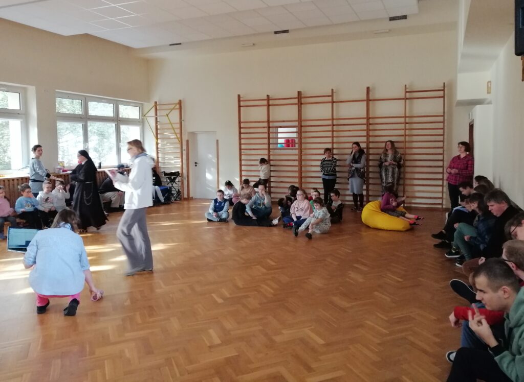 Na sali gimnastycznej siedzą uczniowie szkoły podstawowej. Słuchają utworu muzycznego.