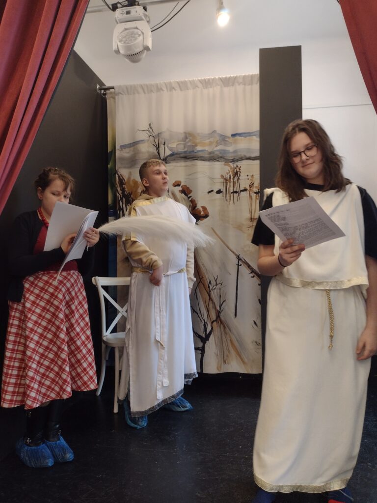 Troje uczniów przebranych w stroje do miniatury dramatycznej czytają teksty trzymane w dłoniach. 