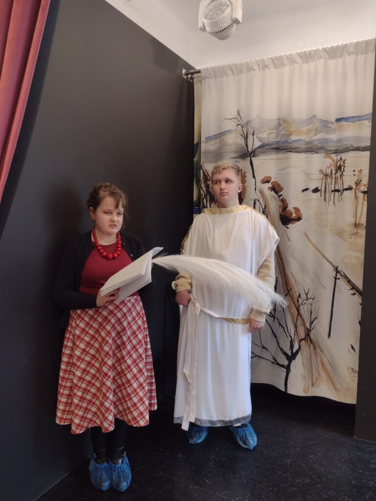 Uczeń wraz uczennicą przebrani w stroje do miniatury dramatycznej. Stoją obok siebie. Dziewczyna czyta tekst miniatury.