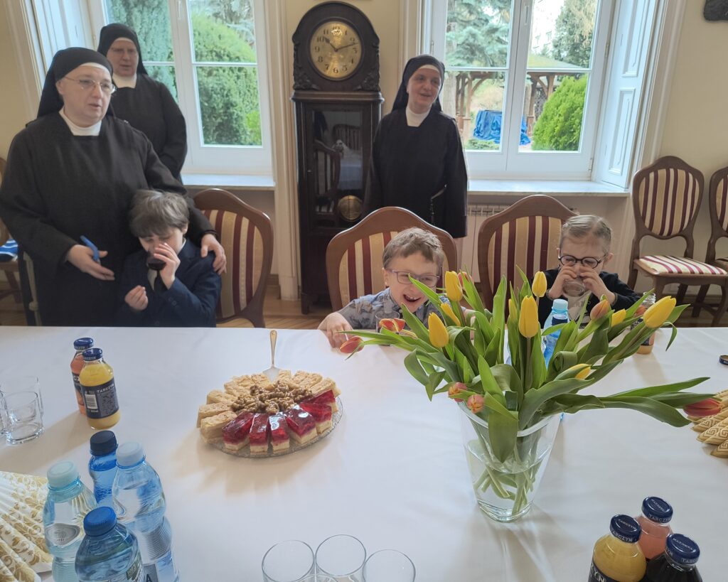 Przedszkolaki siedzą przy stole, za nimi stoją siostry  franciszkanki.