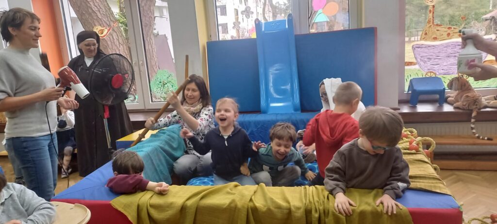 Dzieci w basenie przerobionym na łódź obok pani z suszarką i siostra z wiatrakiem.
