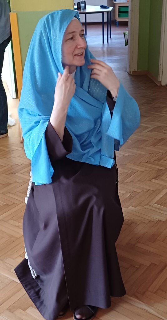 Siostra w niebieskiej chuście na głowie siedzi na krzesełku. 