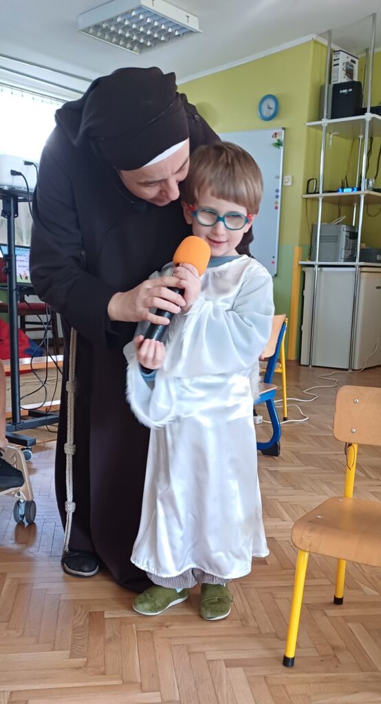 Siostra Kamila pomaga chłopcu w stroju anioła trzymać mikrofon.