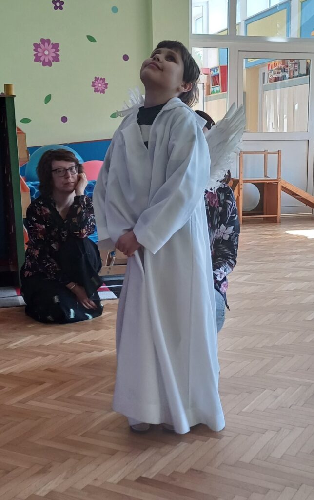 Chłopiec w białym stroju anioła ze skrzydłami na plecach z głową uniesioną do góry. 