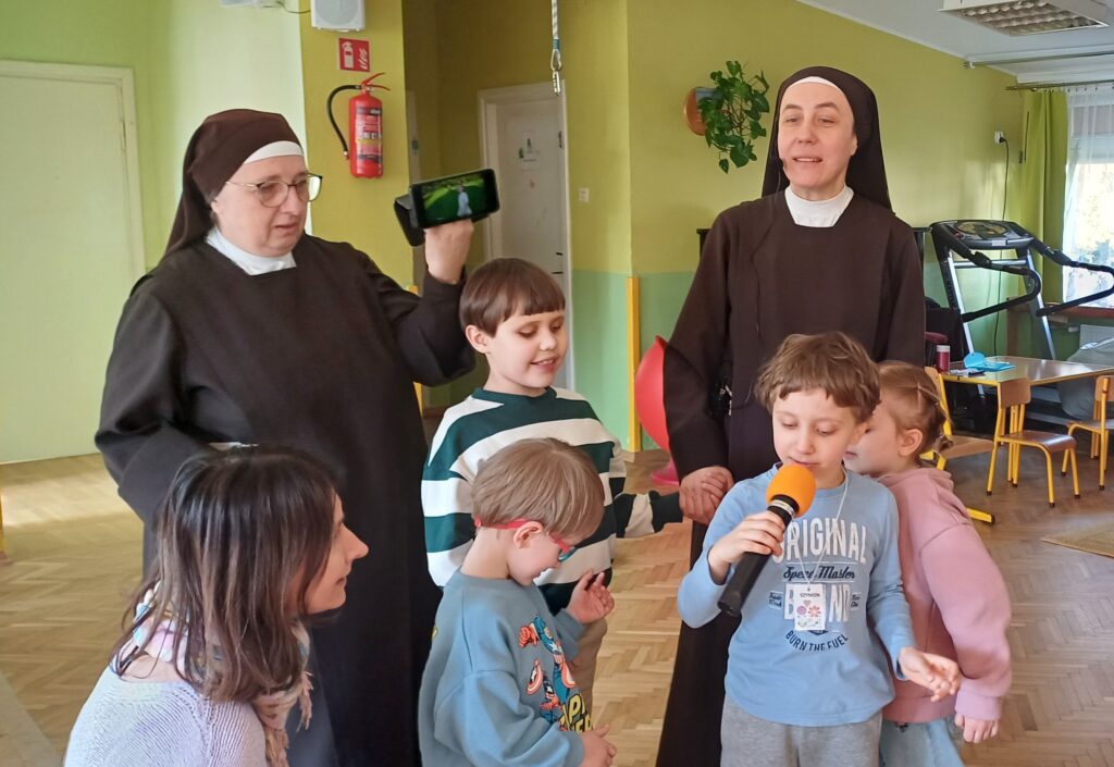 Siostry stoją z dziećmi, jeden chłopiec trzyma mikrofon.