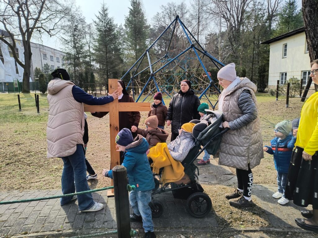 Nauczycielka trzyma stojący krzyż obok dzieci z paniami i siostrą.