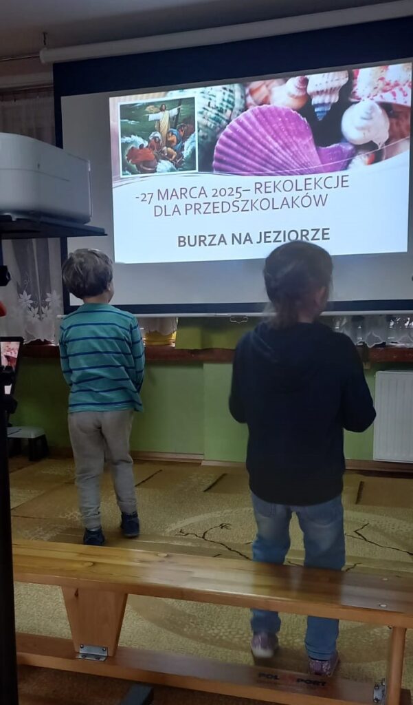 Na dużym ekranie wyświetlony data i temat rekolekcji obok stojące dzieci.