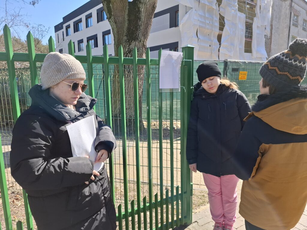 Troje uczniów czyta instrukcje stojąc przy bramie placu zabaw.