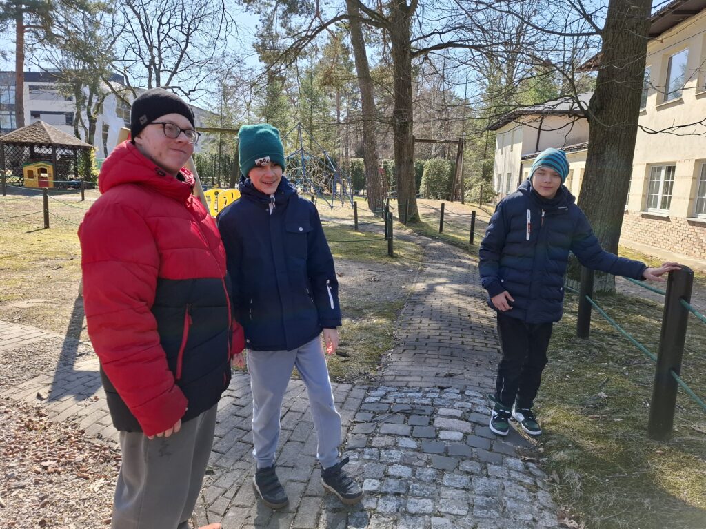 Trzech uczniów uśmiecha się pozując do zdjęcia. Stoją na chodniku przy placu zabaw.