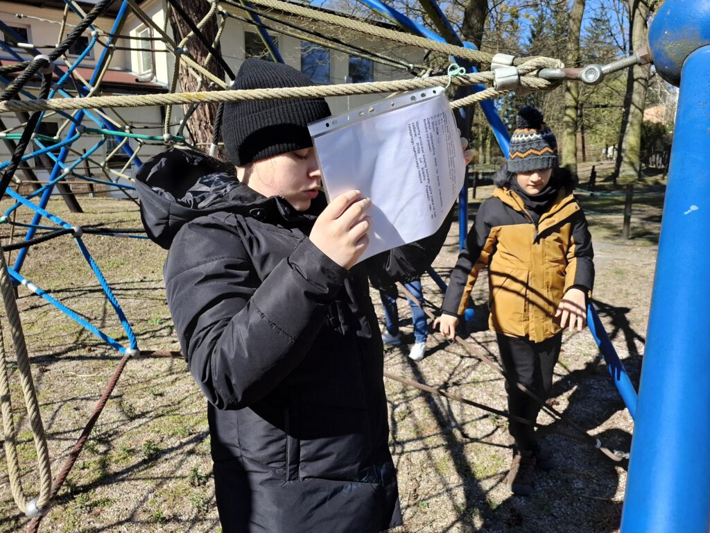 Uczennica czyta instrukcje stojąc obok kolegi na placu zabaw.