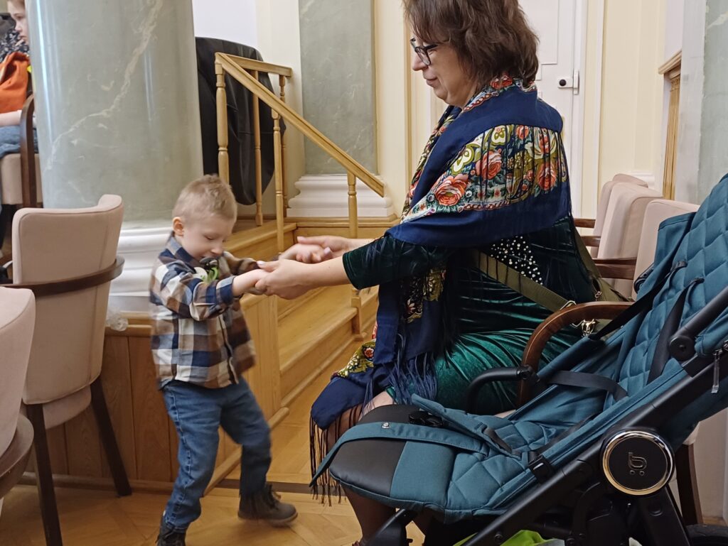 Mały chłopiec stoi a pani nauczycielka trzyma go za ręce.