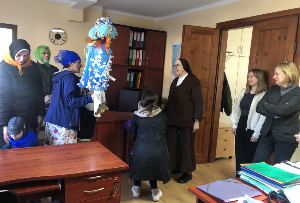Przedszkolaki i nauczyciele w kolorowych chustach z kukiełką Pani Zimy stoją z pracownicami Działu Księgowego.