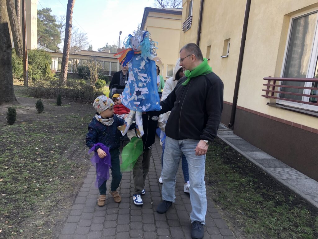Przedszkolaki i nauczyciele z kolorowymi chustami i kukiełką Pani Zimy idą obok Domu Dziewcząt.